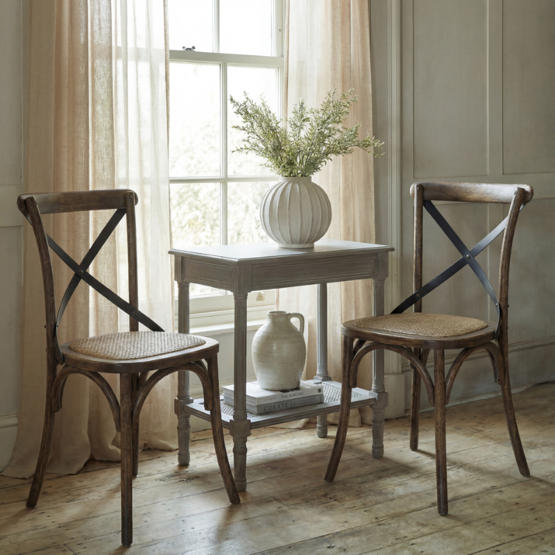 Two rustic wooden dining chairs with woven cane seats and dark cross-back supports, arranged around a small white side table in a bright room with sheer curtains and a window.