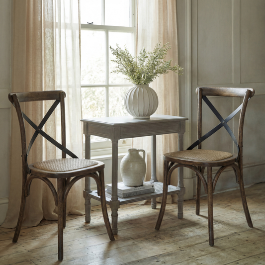 Two rustic wooden dining chairs with woven cane seats and dark cross-back supports, arranged around a small white side table in a bright room with sheer curtains and a window.