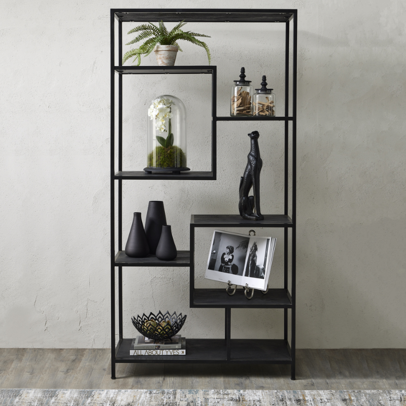 A tall black metal shelving unit with five open shelves, styled with decorative objects including a potted fern, glass cloche, vases, a black figurine, a framed black-and-white photo, and a bowl, set against a textured light gray wall.