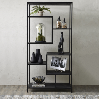 A tall black metal shelving unit with five open shelves, styled with decorative objects including a potted fern, glass cloche, vases, a black figurine, a framed black-and-white photo, and a bowl, set against a textured light gray wall.