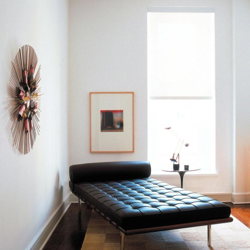 A black Barcelona Pavilion daybed made of genuine leather, positioned in a room with a window, a decorative wall hanging, and a small round table with an object on top.
