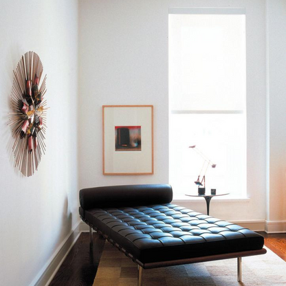 A black Barcelona Pavilion daybed made of genuine leather, positioned in a room with a window, a decorative wall hanging, and a small round table with an object on top.